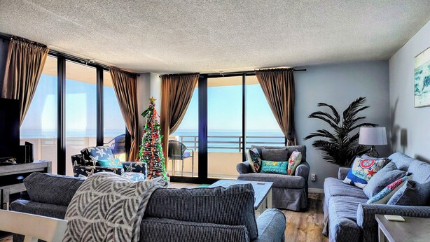 A living room with a view of the ocean, a Christmas tree, and a balcony with a table and chairs. - Roomy Oceanfront 2/2 condo with Million Dollar views in fabulous Daytona Beach!! (Daytona Beach)