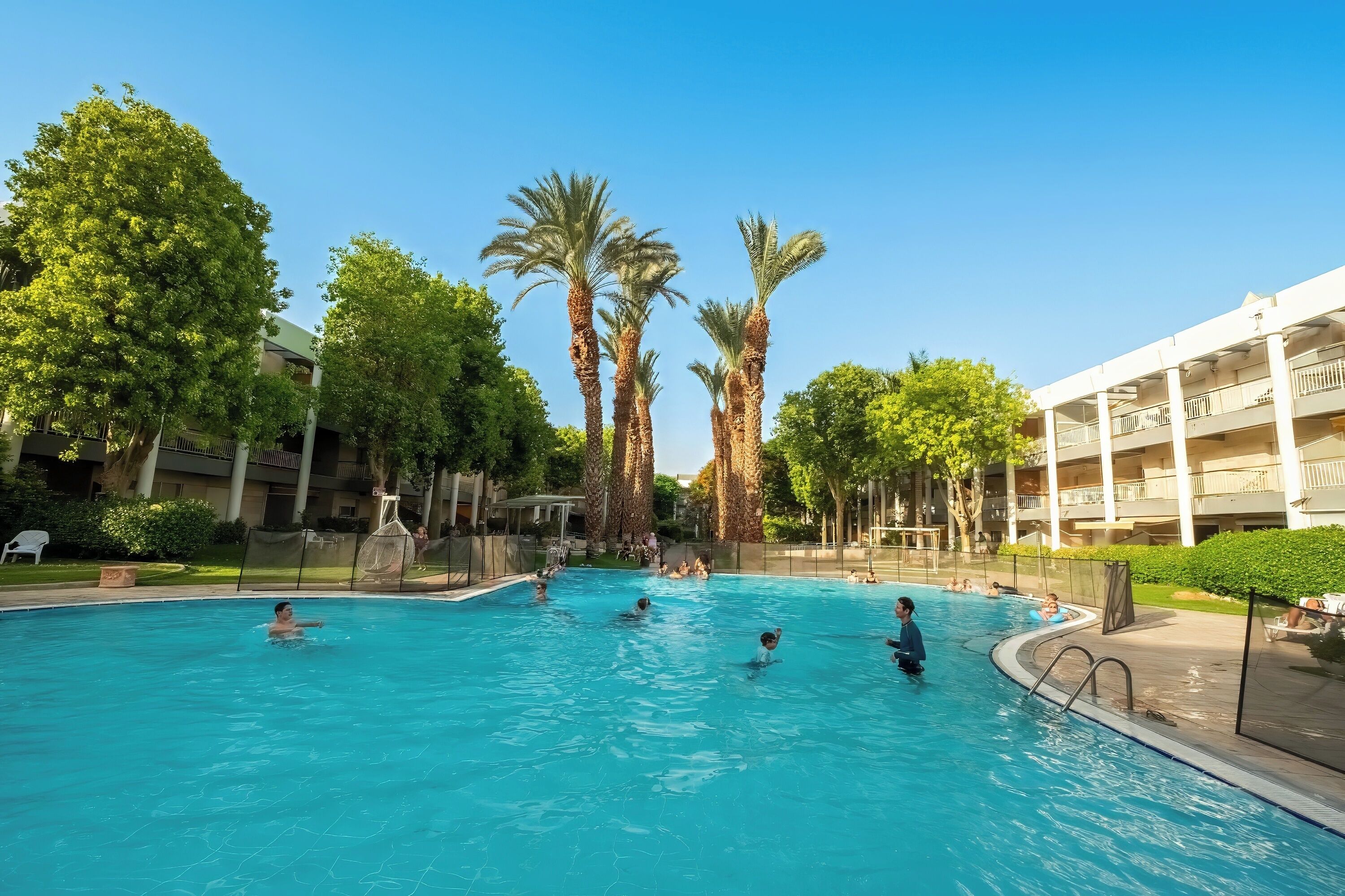 A swimming pool surrounded by palm trees and a multi-story building with balconies.