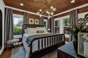 A bedroom with a wooden ceiling, a bed with a dark headboard, a bench, and a potted plant.