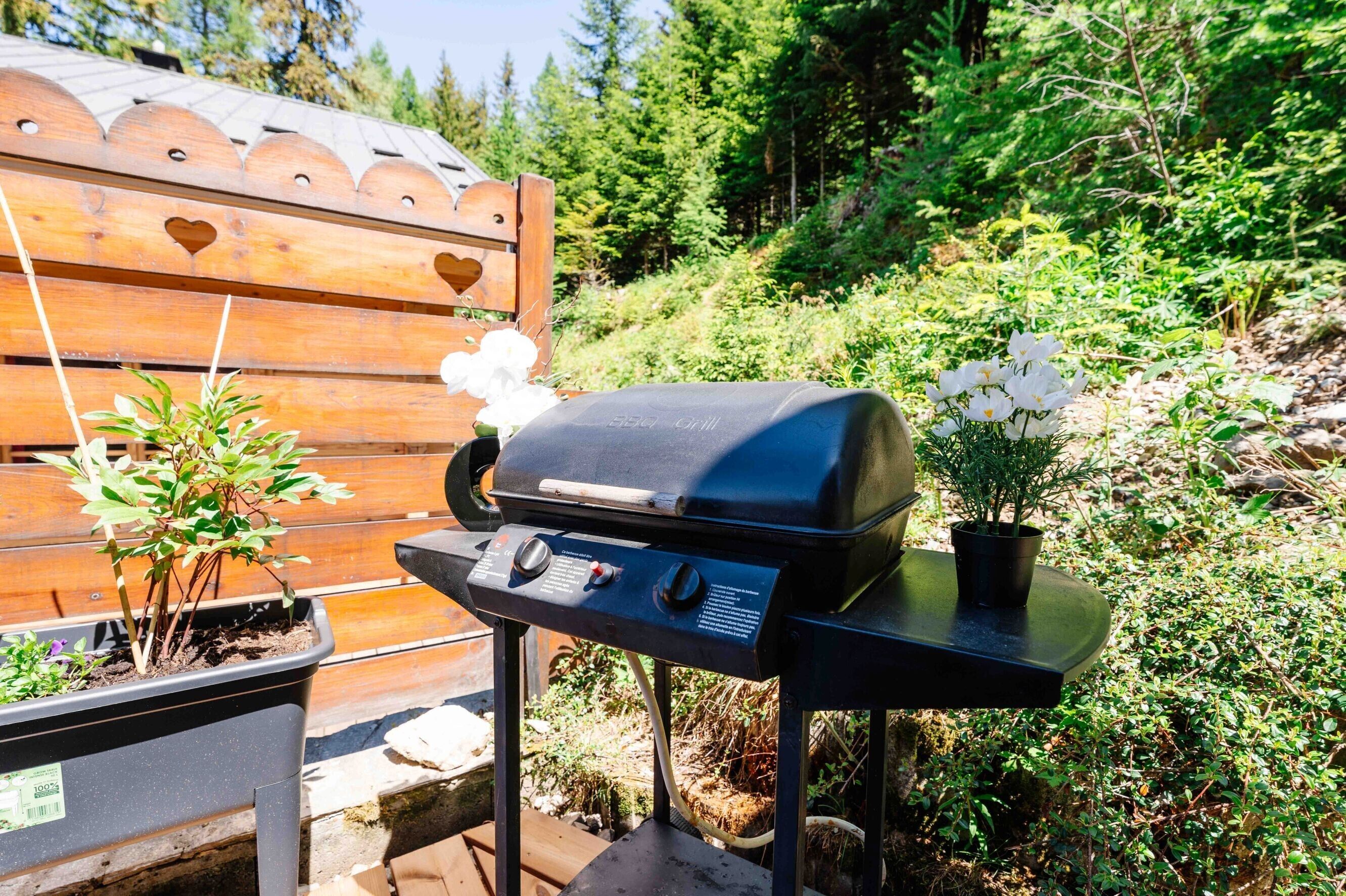 A portable grill with control knobs, a potted plant, and a wooden bench in a garden setting.