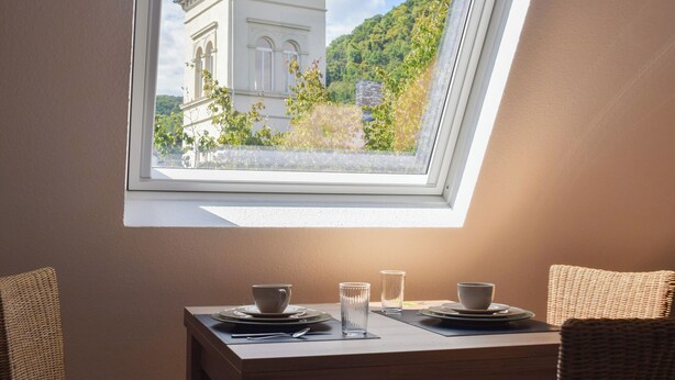 A dining area with a table set for two, a window with a view of a building and greenery, and wicker chairs.