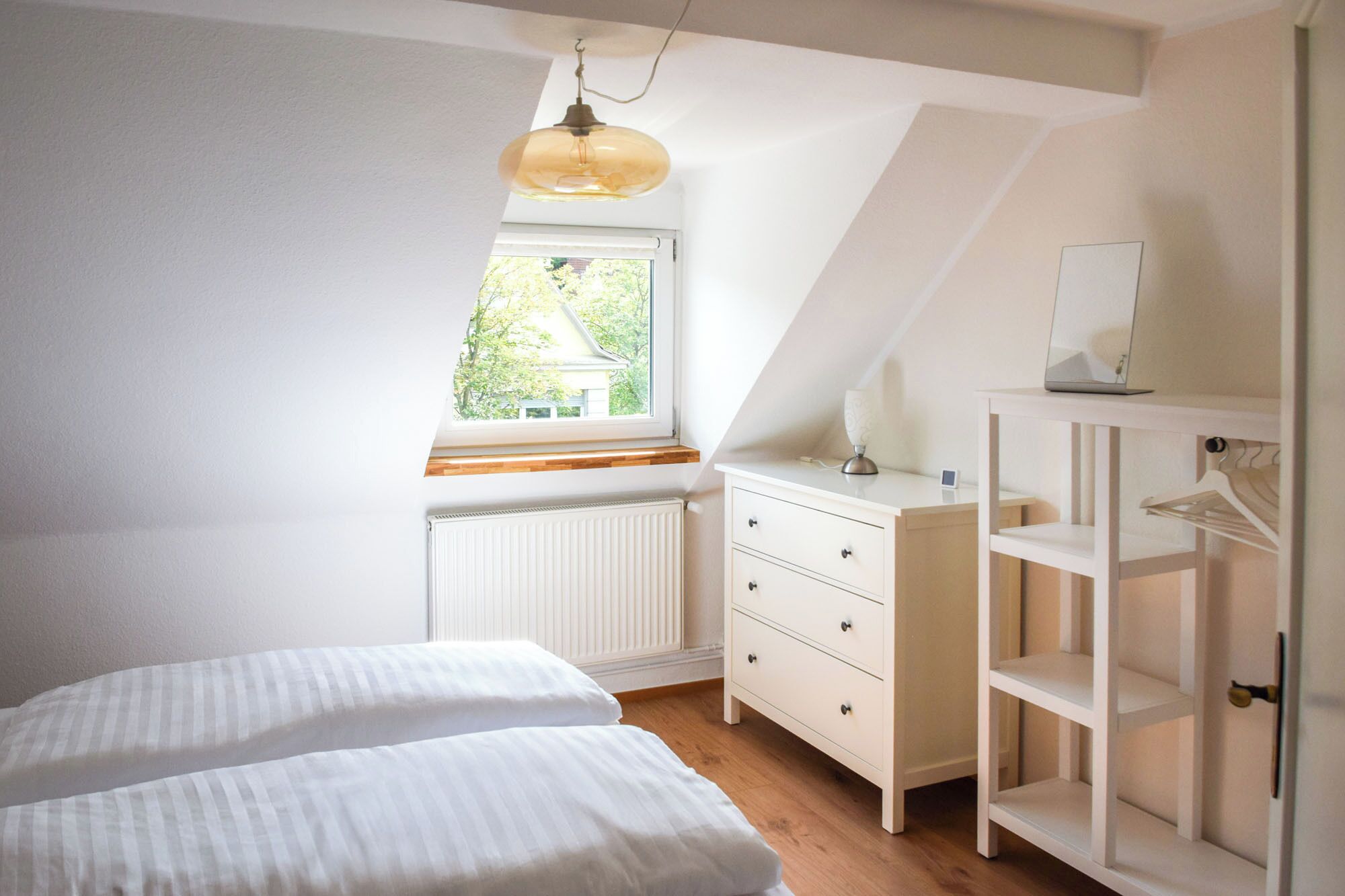 A bedroom with a sloped ceiling, a window with a view of greenery, a white dresser with drawers, a bedside table with a lamp, and a bed with white bedding.
