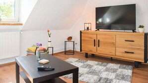 A cozy living room with a wooden cabinet, a flat-screen TV, a small table with a remote control, and a window providing natural light.