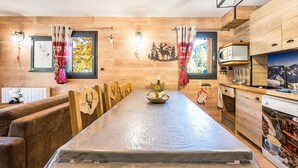 A wooden dining table with chairs, a kitchen area with a microwave, and a view of the outdoors through a window. - Apartment 'Chalet Refuge Loup Blanc Valfréjus' with Mountain View, Balcony and Wi-Fi (Valfréjus)