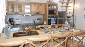 Cocina con armarios de madera, microondas y refrigerador. Se observa una mesa de comedor con sillas y una lámpara colgante.
