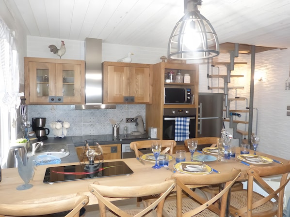 Une cuisine avec des meubles en bois, un four à micro-ondes et un réfrigérateur. On y trouve une table à manger avec des chaises et un luminaire suspendu.