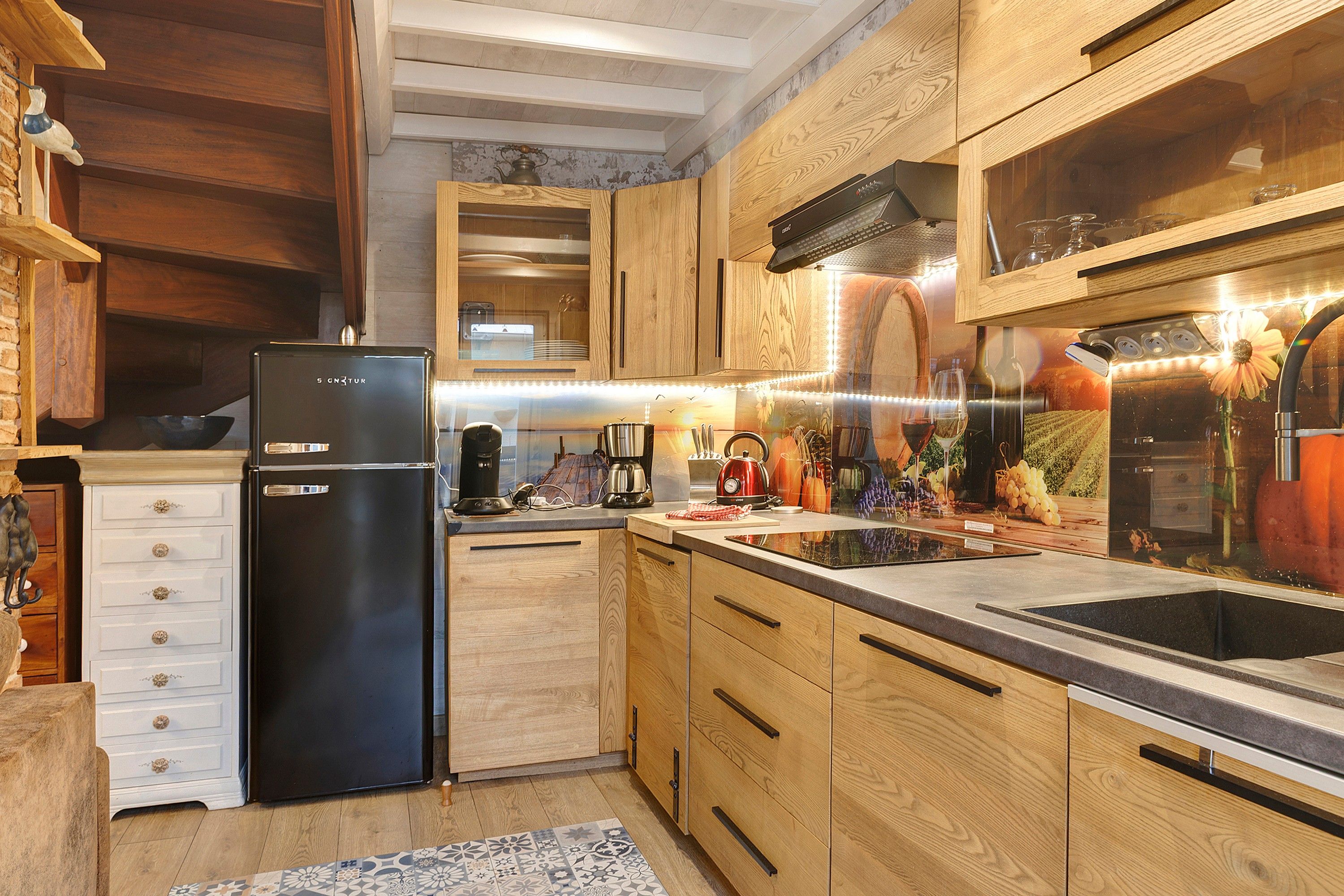 Eine moderne Küche mit Holzschränken, einem schwarzen Kühlschrank und einem großen Fenster mit Blick auf einen Weinberg.