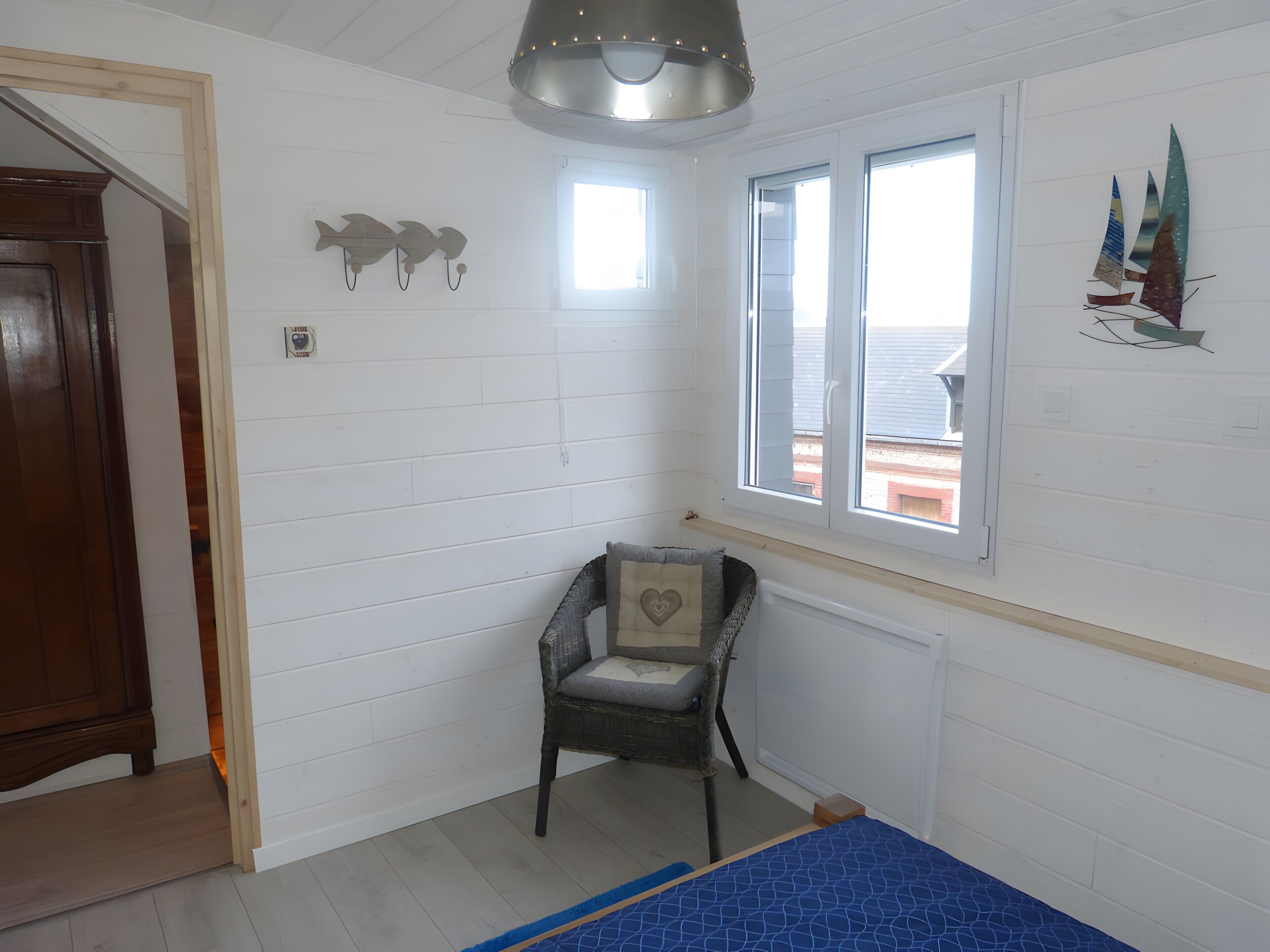 Una habitación con paredes blancas empañadas, una ventana con vistas, una silla con cojín y una cama con cubrecama azul.