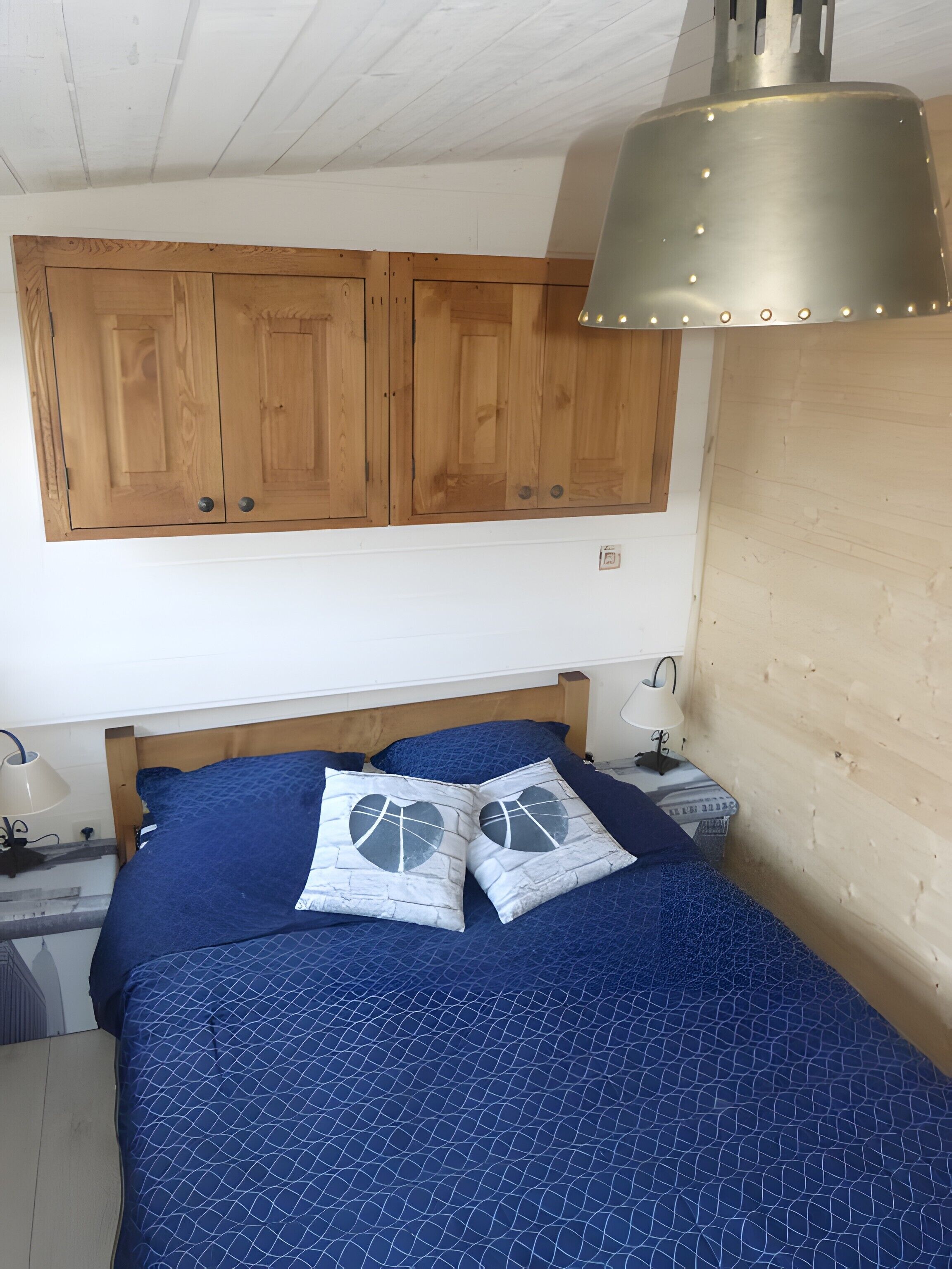 A bedroom with a wooden headboard, a bed with blue bedding, two bedside lamps, and wooden cabinets above the bed.
