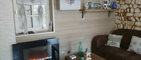 A living room with a fireplace, a television, a stone wall, a wooden shelf with decorative items, and a brown leather sofa.