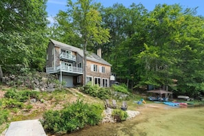 A two-story log cabin with a balcony, surrounded by dense forest and a small dock with kayaks. - Merrymeeting Lake Private Beach, Gazebo & Hot Tub (New Durham)
