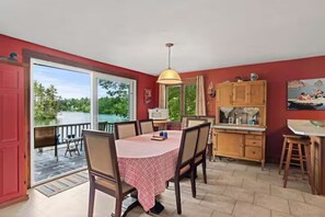 A dining area with a table and chairs, a kitchen with wooden cabinets, and a view of a lake through large windows. - Merrymeeting Lake Private Beach, Gazebo & Hot Tub (New Durham)
