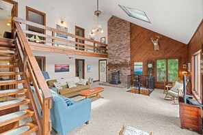 A spacious living area with a staircase, a fireplace, and a large window overlooking greenery. - Merrymeeting Lake Private Beach, Gazebo & Hot Tub (New Durham)