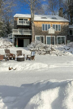 A two-story house with a covered deck, a garage, and a snow-covered yard with outdoor seating. - Merrymeeting Lake Private Beach, Gazebo & Hot Tub (New Durham)