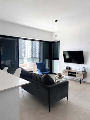 Un salon moderne avec un canapé, un fauteuil bleu, un téléviseur et une table basse en verre.