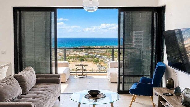 A modern living room with a sofa, chairs, a coffee table, a TV, and a balcony with a view of the ocean. - Elegant Tower Apt | Sea View & Parking | Mamad (Bat Yam)