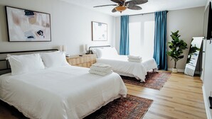 A hotel room with two beds, a ceiling fan, a large window with blue curtains, a plant, and a mirror. - Convenient 2BR Escape | Easy Walk to MOB (Mobile)