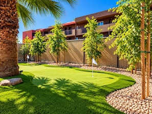 A well-maintained golf course with a putting green, surrounded by a fence, trees, and a building with multiple balconies.