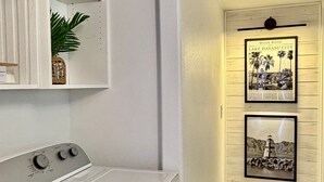 A laundry room with a white washing machine, a shelf with decorative items, and a door with framed pictures. - Endless Summer, Pool/Theatre/Boat Parking/Walk to London Bridge! (Lake Havasu City)