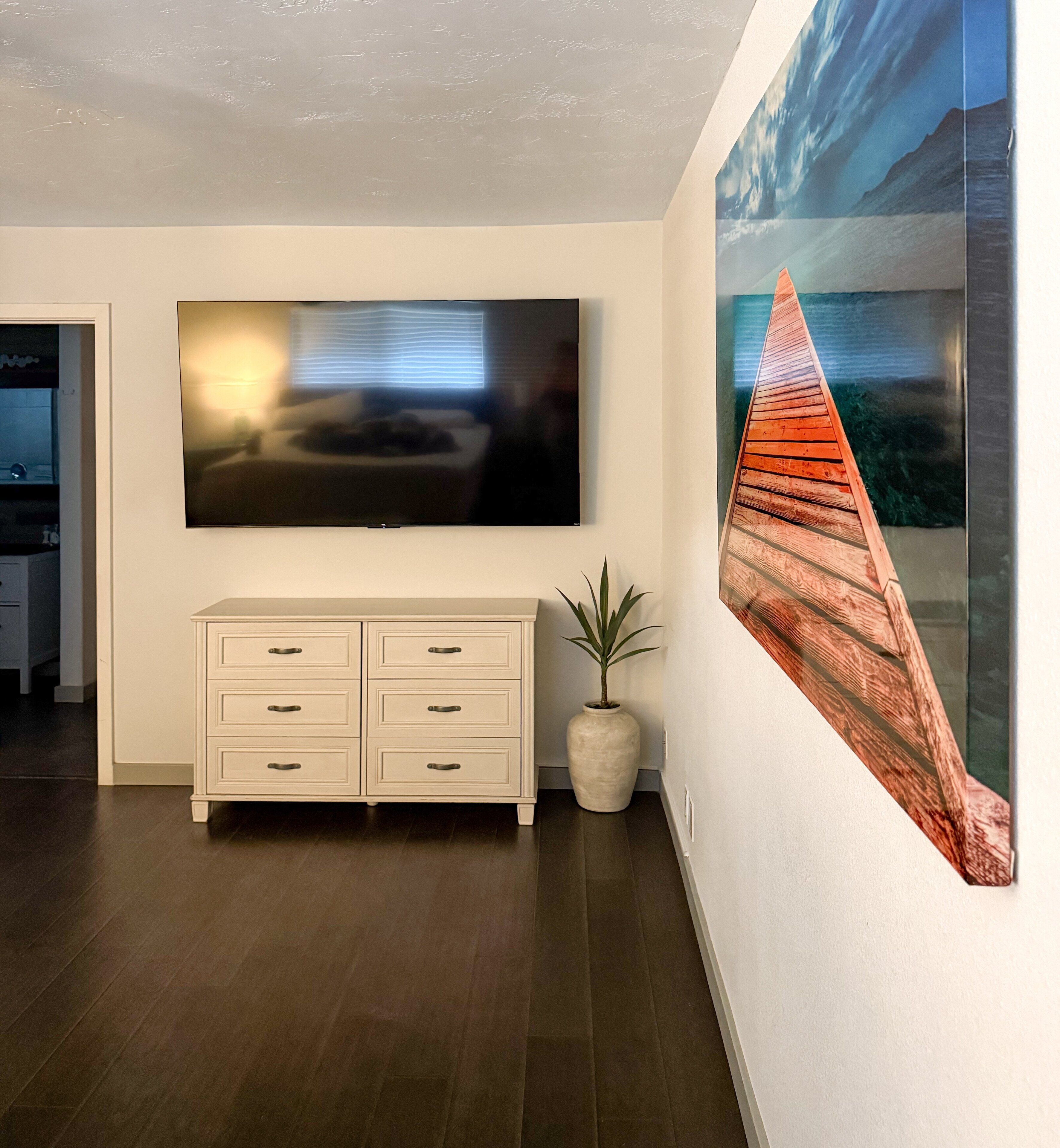 Un salon moderne avec un téléviseur à écran plat fixé au mur, un meuble blanc avec des tiroirs et un grand tableau représentant un bateau en bois, également accroché au mur.