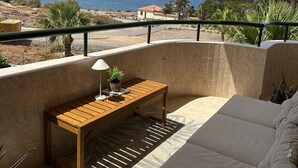 A balcony with a wooden table, a sofa, and a view of the sea and a mountain.
