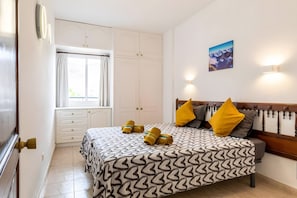 A bedroom with a bed, a window with curtains, a wardrobe, a painting on the wall, and a wall-mounted light.