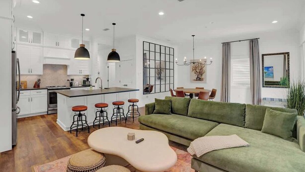 A modern kitchen with white cabinets, a central island with stools, pendant lighting, and a green sofa in the living area. - Church St Collection - Suite 16 (Fairhope)