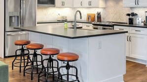 A modern kitchen with white cabinets, stainless steel appliances, a central island with stools, and pendant lighting. - Church St Collection - Suite 16 (Fairhope)
