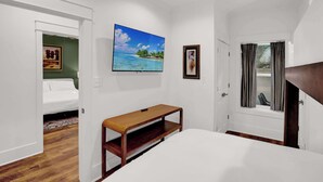 A bedroom with a bed, a wooden nightstand, a TV mounted on the wall, and a window with curtains. - Church St Collection - Suite 16 (Fairhope)