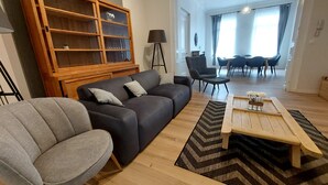 Un salon avec un canapé gris, un meuble en bois, un fauteuil gris, une table basse et, au fond, un coin repas.