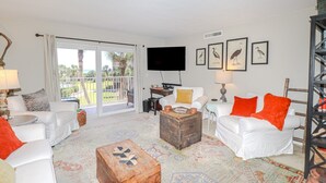A spacious living room with white furniture, a wooden coffee table, and a large window with a view of palm trees. - New to Rental Program! Beautiful Ocean View 3/2 at St. Augustine Beach & Tennis Club 301 (St. Augustine)