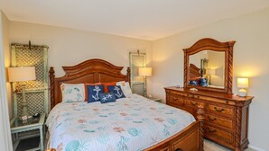 A bedroom with a wooden bed, two bedside tables with lamps, a dresser with a mirror, and a floral bedspread. - New to Rental Program! Beautiful Ocean View 3/2 at St. Augustine Beach & Tennis Club 301 (St. Augustine)