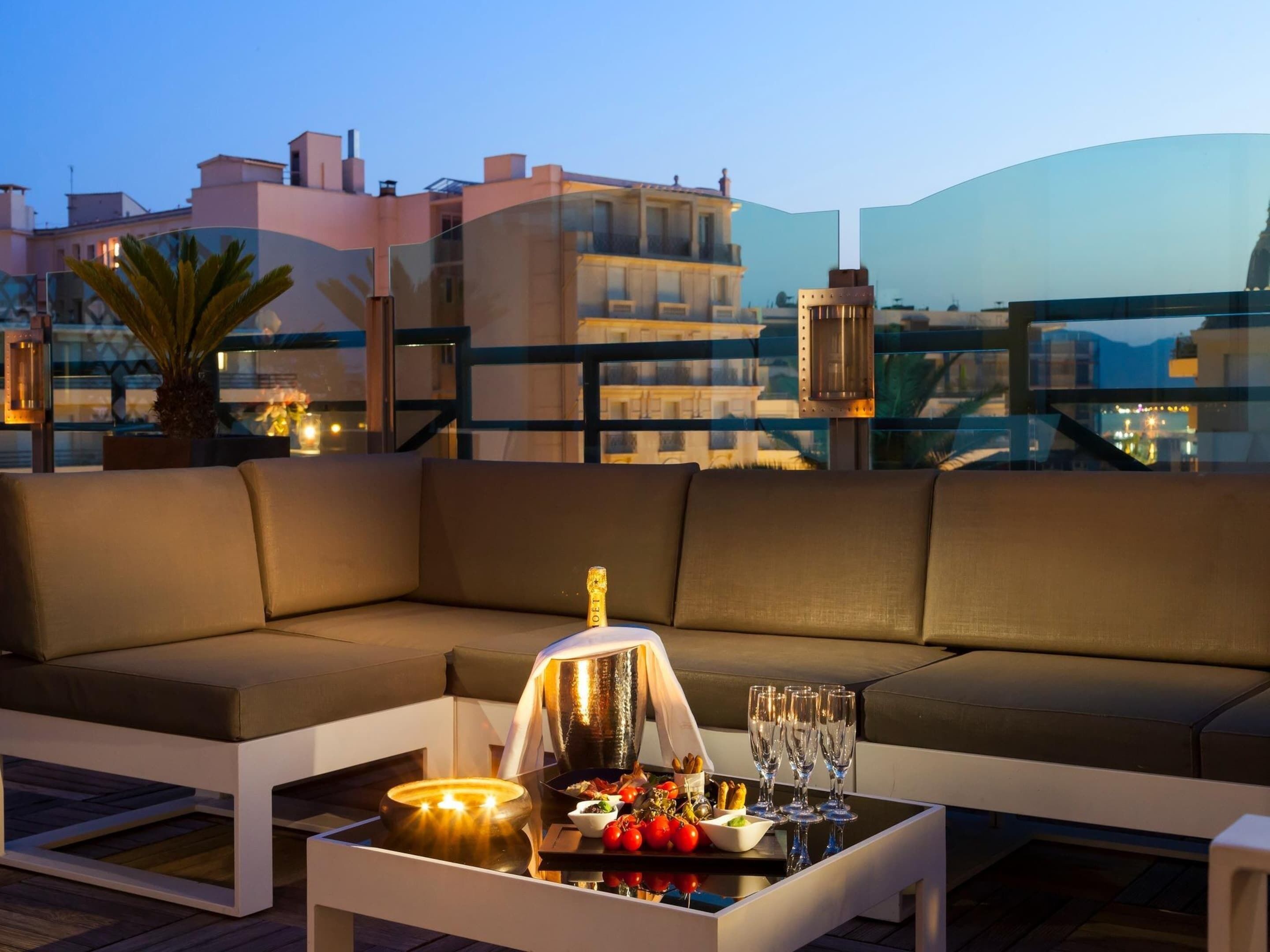 A rooftop terrace with a sofa, a table with a bottle, glasses, and food, overlooking a cityscape at dusk.