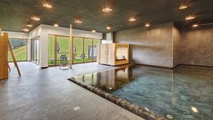 Un área de spa con una amplia piscina interior, bancos de madera y vistas a un paisaje con césped a través de grandes ventanales.