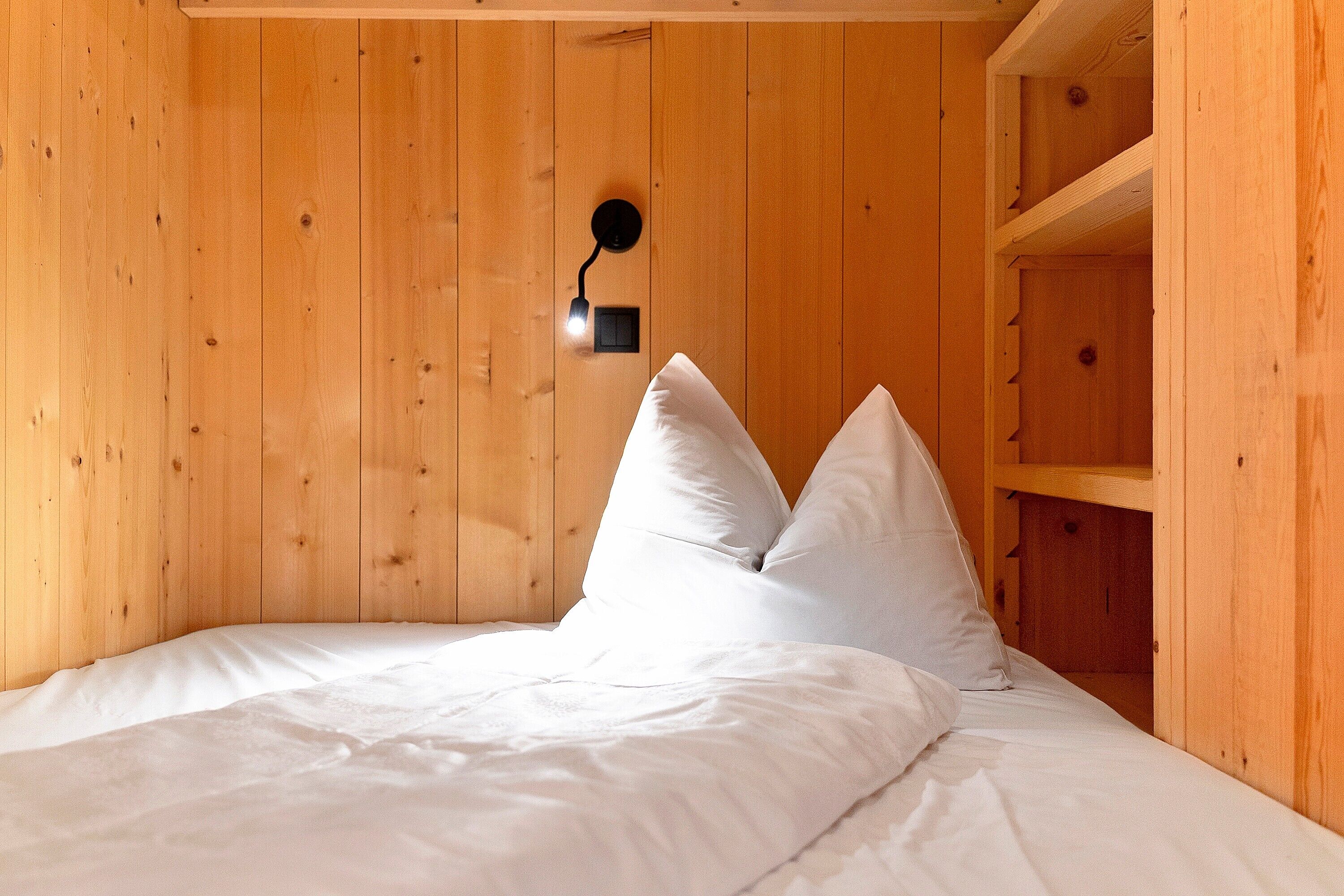 Una habitación de madera con una cama, ropa de cama blanca y una lámpara fijada a la pared.