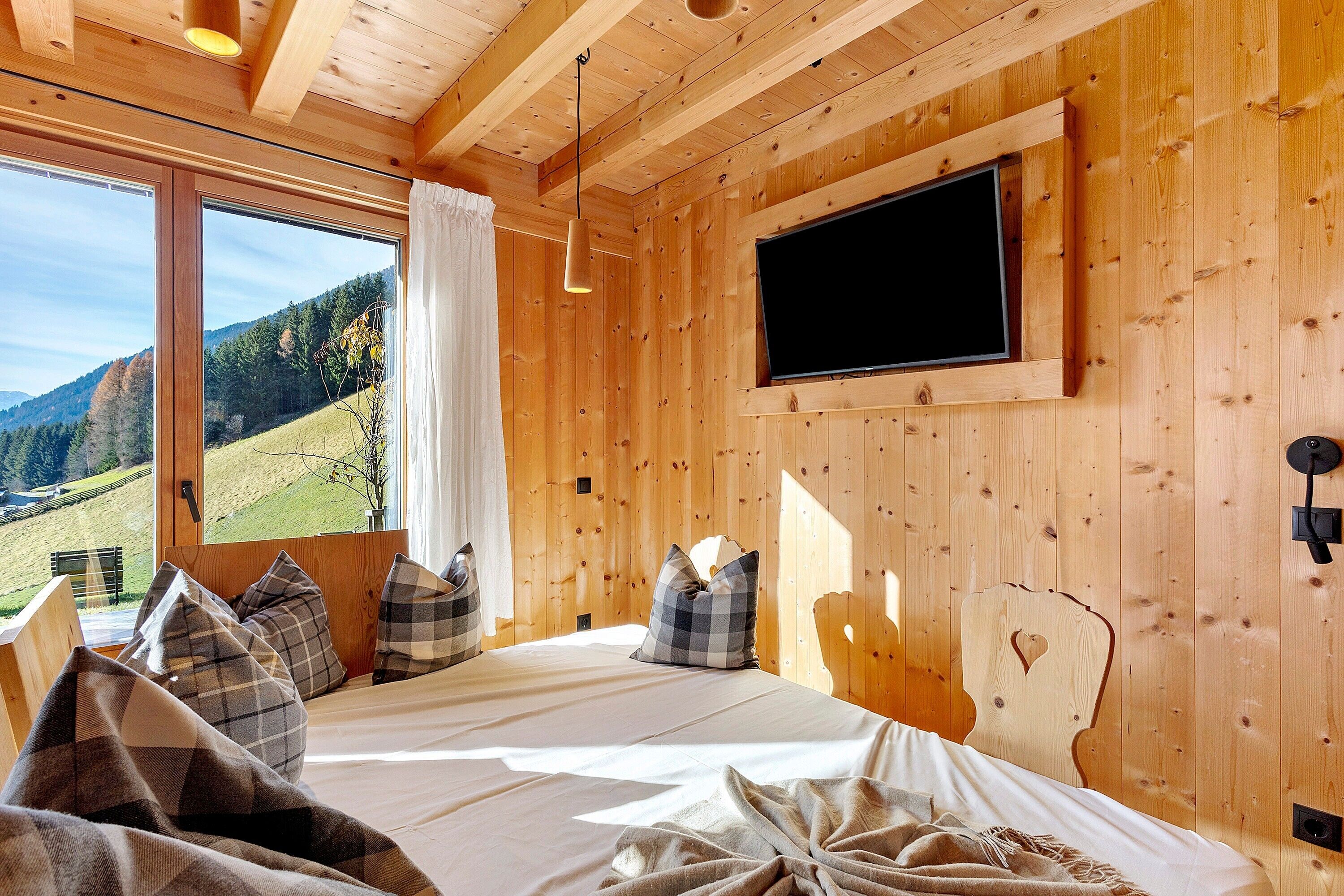 Una habitación tipo cabaña de madera con una cama, un televisor montado y una ventana con vistas al paisaje.