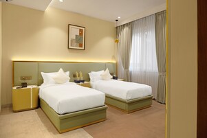 Quarto de hotel com duas camas, uma mesa de cabeceira, uma luminária, um quadro na parede e uma janela com cortinas.