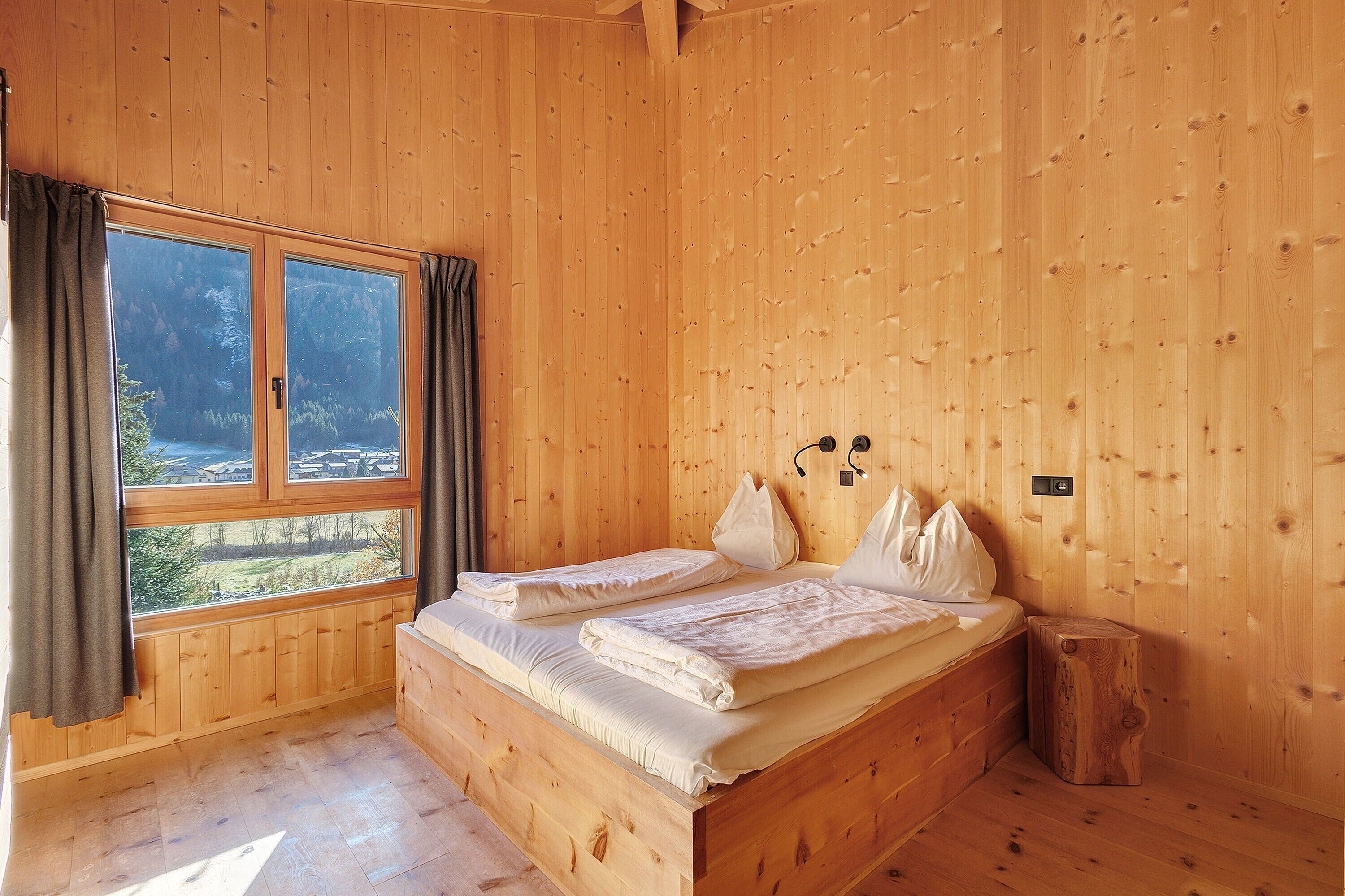 Una habitación de madera con una cama, un ventanal y un taburete de madera.