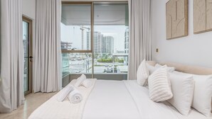 A modern hotel room with a large bed, white linens, and a view of a cityscape through floor-to-ceiling windows. - Ellington Beach House - Palm Jumeirah (Dubai)