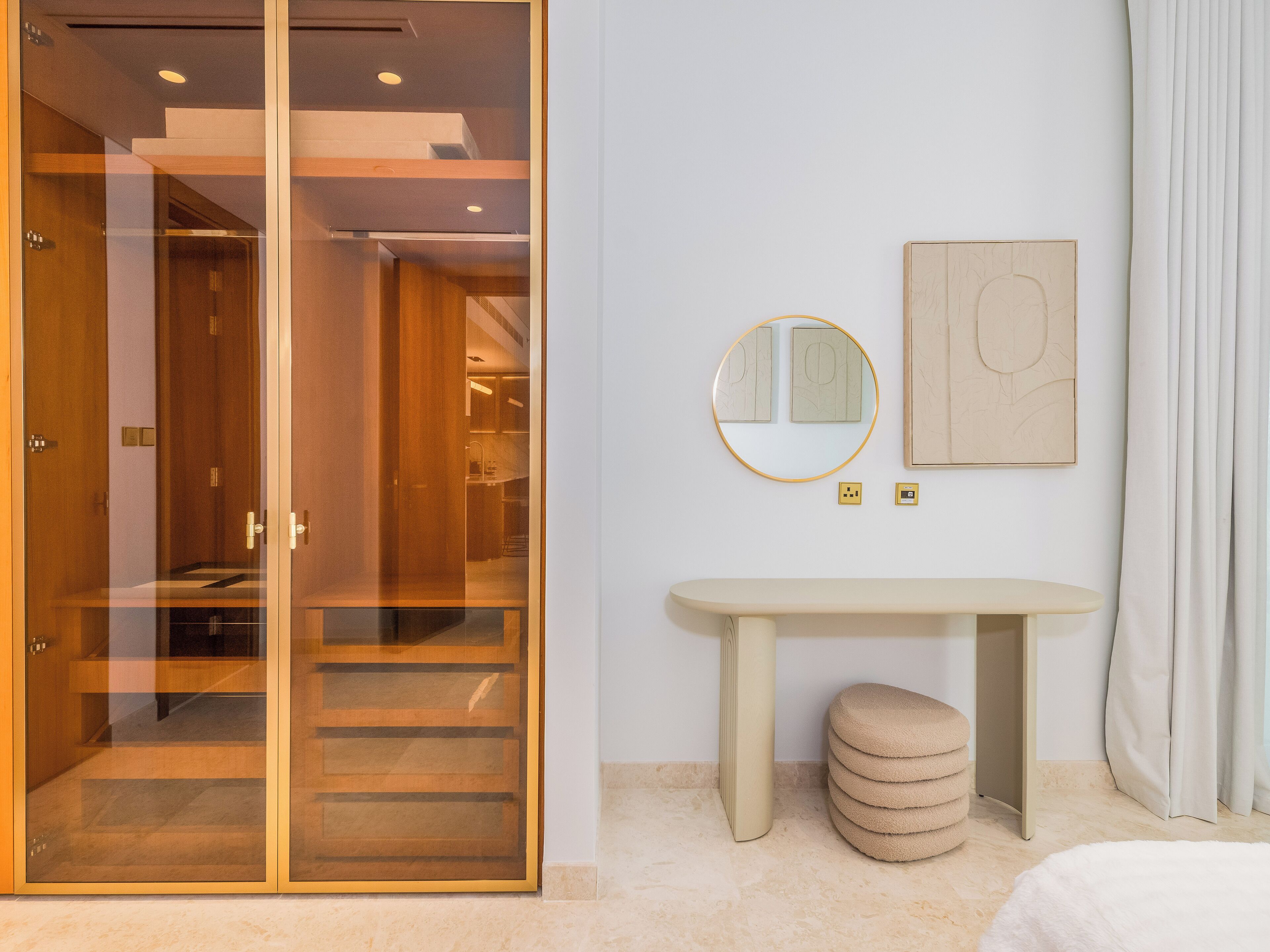 A modern hotel room with a glass door leading to a hallway, a round mirror, a small table, and a round ottoman.