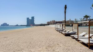 A beach with sun loungers and a pool area, a clear sky, and modern buildings in the background. - Ellington Beach House - Palm Jumeirah (Dubai)