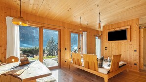 L’intérieur d’un chalet en bois comprend une table à manger, des chaises, un lit avec une tête de lit, un téléviseur fixé au mur et de grandes fenêtres offrant une vue sur l’extérieur.