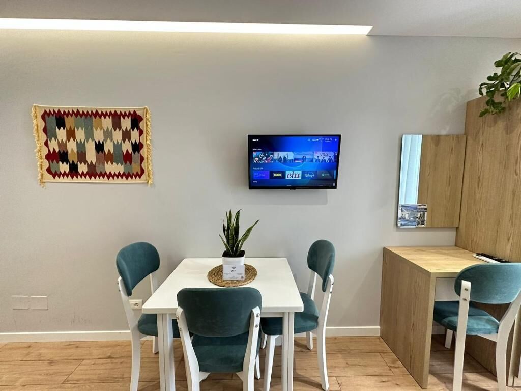 Een moderne eetruimte met een witte tafel, teal stoelen, een kleine potplant en een flatscreen televisie aan de muur.