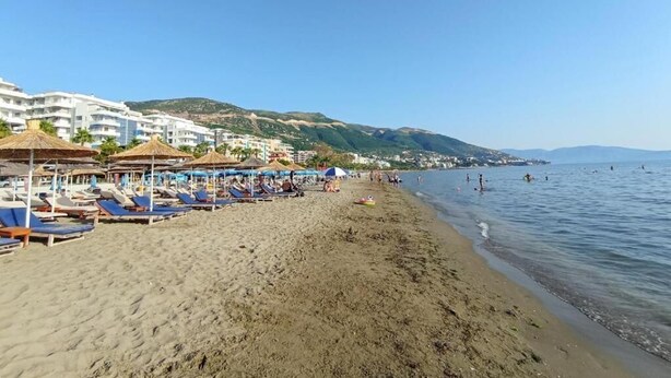 Ein Strand mit Liegestühlen und Sonnenschirmen, einem klaren Himmel und einer hügeligen Landschaft im Hintergrund.
