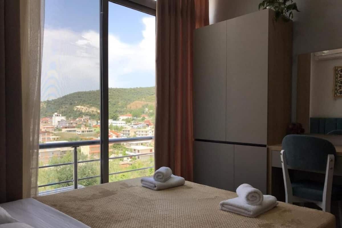 A hotel room with a large window, a bed, a desk, and two rolled towels on the bed.