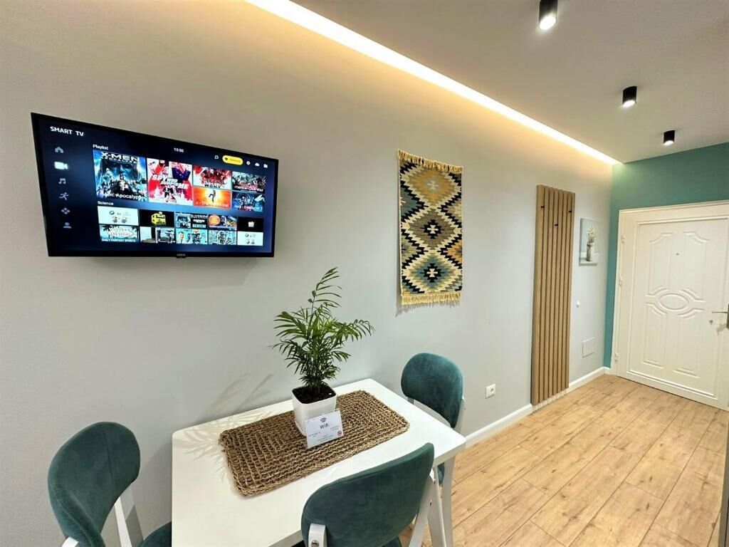 Un salon moderne avec un téléviseur à écran plat fixé au mur, qui affiche l’interface d’un service de diffusion en continu. On y trouve une petite table sur laquelle est posée une plante en pot et un set de table en fibres tressées. La pièce est dotée d’un plancher en bois et une porte est visible au fond.