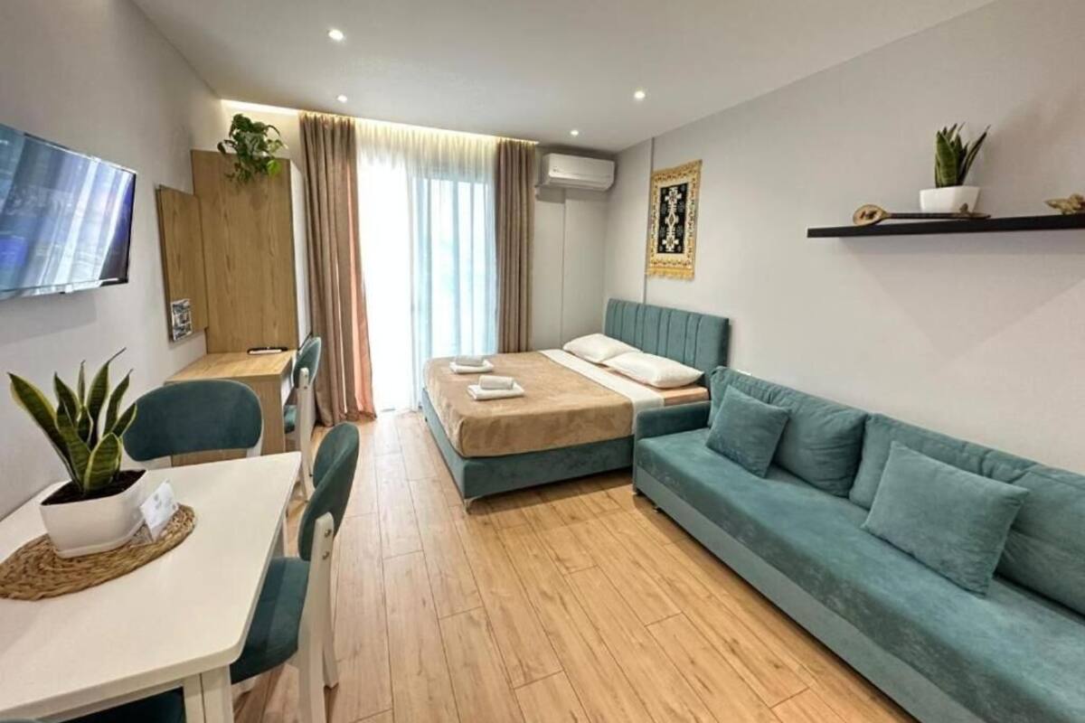 A modern hotel room with a bed, a sofa, a dining area with a table and chairs, a TV, and a potted plant.