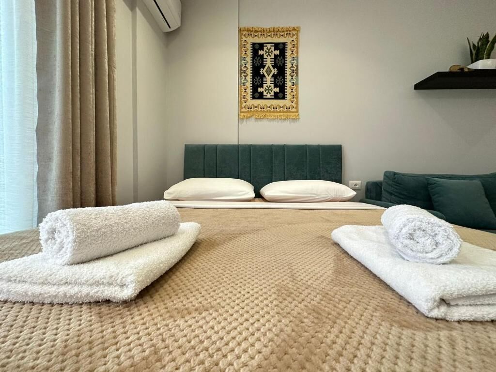 A hotel room with a bed, two pillows, a wall-mounted air conditioner, a decorative tapestry, and a small plant on a shelf.