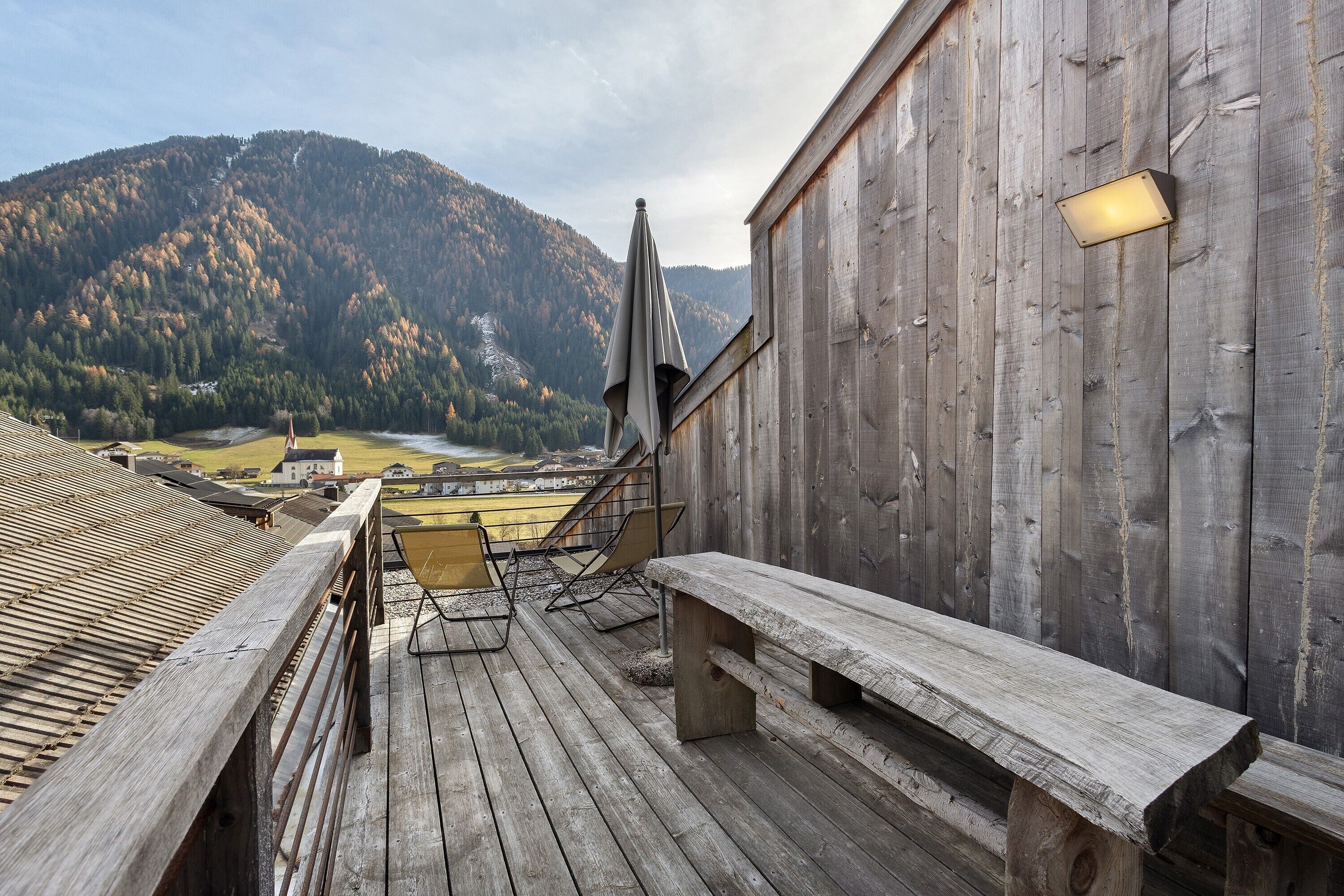 A wooden deck with a bench, a chair, and a light fixture on a building.