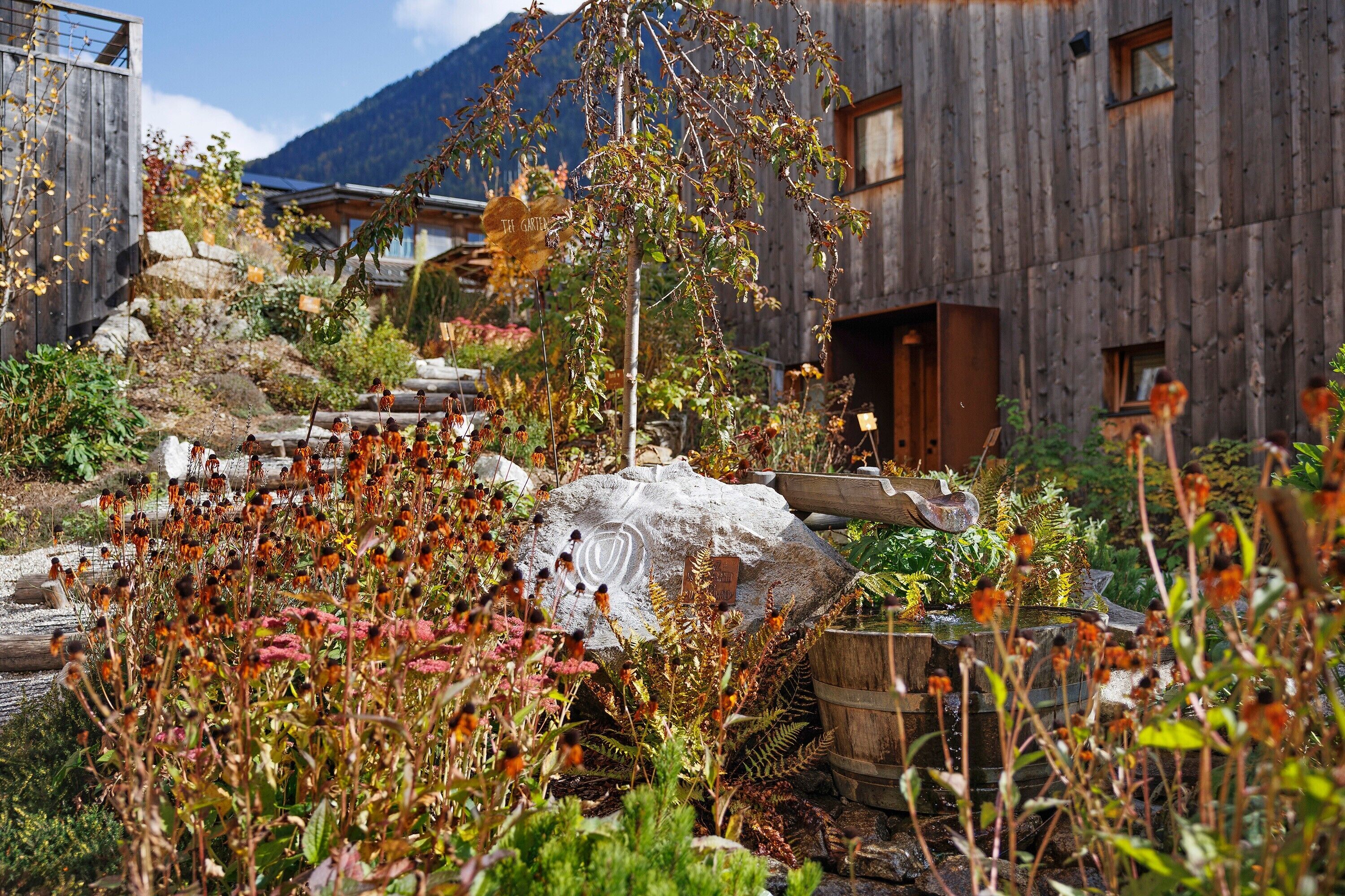 A rustic wooden building with a garden in front, a wooden staircase, and a mountain in the background.